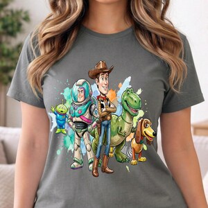 May include: A charcoal gray short-sleeve t-shirt featuring a colorful graphic print of characters from Toy Story, including a green alien, a space ranger, a cowboy, a green dinosaur, and a dachshund toy, set against a watercolor splash background.