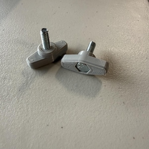 Puede incluir: Dos perillas en forma de T de color gris claro con pernos metálicos integrados. Cada perilla presenta un diseño de mango alargado y texturizado con una cabeza de perno hexagonal de metal incrustada en el plástico para un agarre manual.