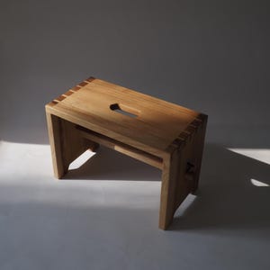 Könnte beinhalten: Ein hellfarbener Holztritt mit rechteckiger Oberseite und einem Griff. Der Hocker hat vier Beine und einen horizontalen Stützbalken. Die Holzmaserung ist sichtbar. Der Hocker steht vor neutralem Hintergrund.