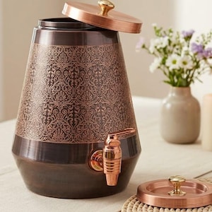 May include: A tapered copper-colored beverage dispenser with a dark brown finish and an intricate embossed floral pattern around the upper body. It features a matching copper-colored spigot and a removable lid with a small knob handle on top.