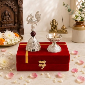 May include: A silver-toned ritual bell with a bird-shaped handle and a brown rudraksha bead, paired with a silver-toned oil lamp containing a lit flame, both resting on a rectangular red velvet box with a gold ribbon and a gold metal latch.
