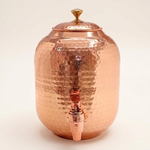 May include: A hammered copper water dispenser with a cylindrical body, a removable lid featuring a small brass-colored knob, and a matching metallic spigot attached to the lower front section. The surface has a reflective, textured finish.