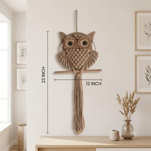 May include: A tan macrame owl wall hanging featuring dark brown eyes, a textured knotted body, and long fringe tail feathers. The owl is perched on a wooden dowel and measures 23 inches in height by 12 inches in width as indicated by the text.