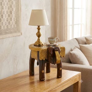 Puede incluir: Una mesa auxiliar de madera marrón oscuro con forma de elefante y detalles en metal dorado en las orejas, la trompa y las patas. Una lámpara de mesa de latón con pantalla color crema y una pequeña taza de cerámica descansan sobre la superficie.