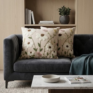 May include: Two square throw pillows featuring a light beige fabric base with embroidered white daisy flowers, brown centers, green stems, and small pink accents. The floral design is textured and covers the front of each pillow.