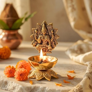 Peut inclure: Une lampe à huile en laiton doré avec une figure de divinité, allumée avec une flamme. La lampe est sur une base de fleur de lotus, avec des fleurs et des pétales orange à proximité. L'arrière-plan comprend un pot en cuivre et un tissu drapé.