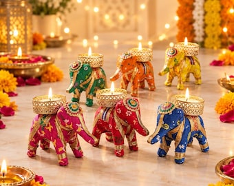 Op de afbeelding: Een verzameling van zes olifantvormige kaarsenhouders in levendige kleuren zoals roze, groen, oranje, geel, rood en blauw. Elke olifant heeft een goudkleurige metalen theelichthouder op de rug met een brandend waxinelichtje.
