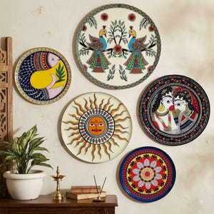 May include: Five circular wall plates featuring vibrant hand-painted designs including two peacocks, a stylised sun, a fish, a floral mandala, and two figures. The plates are arranged in a cluster, showcasing intricate patterns in red, blue, yellow, and black.