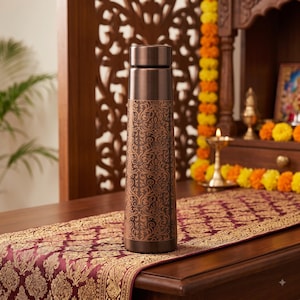 Peut inclure: Une bouteille d'eau cylindrique en métal avec une finition marron foncé et un motif floral répétitif de couleur cuivre couvrant les deux tiers inférieurs du corps. La bouteille présente un bouchon lisse et uni marron foncé.