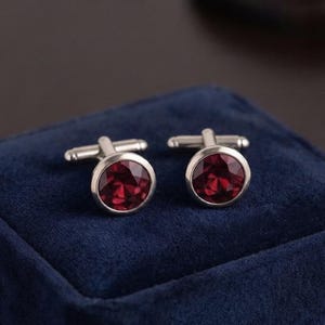 May include: A pair of round cufflinks featuring deep red faceted stones set in a polished silver-toned metal frame. Each cufflink has a classic T-bar swivel mechanism on the back, designed to secure through shirt cuffs with a circular front face.