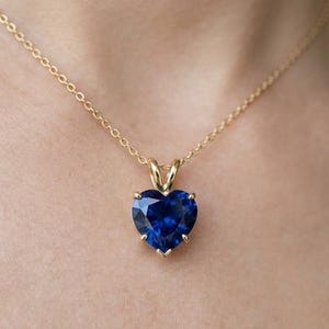 May include: A gold necklace featuring a heart-shaped, deep blue gemstone pendant. The pendant is set in a gold bezel and suspended from a delicate gold chain. The necklace is shown against a neutral background.