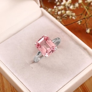 Puede incluir: Un anillo plateado con una gran gema rosa claro de forma rectangular. La gema está engastada en un engaste de cuatro puntas. El anillo se presenta en una caja de joyería blanca.