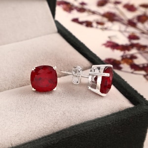 Könnte beinhalten: Ein Paar rubinrote Edelstein-Ohrstecker mit Facettenschliff, gefasst in Silbermetall. Die Ohrringe werden in einer dunkelgrünen Schmuckschatulle mit hellbeigem Innenraum präsentiert. Die Edelsteine haben eine quadratische Form.