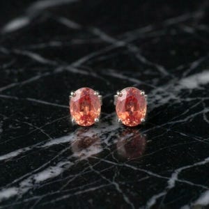 Puede incluir: Un par de pendientes de botón con piedras preciosas de color melocotón de corte ovalado. Las piedras están engastadas en un metal plateado. Los pendientes se exhiben sobre una superficie de mármol blanco y negro.