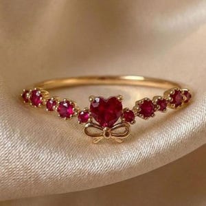 May include: A gold ring featuring a central heart-shaped red gemstone accented by a small gold bow underneath. The band is adorned with a series of smaller round red gemstones extending outward from the center on both sides of the ring.