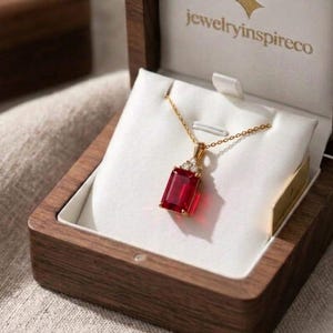 May include: A gold necklace featuring a rectangular red gemstone pendant with small clear stones at the top, displayed in an open dark wood jewelry box with the text jewelryinspireco printed in gold on the white interior lining.