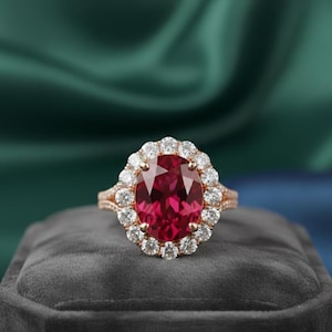 Può includere: Un anello in oro rosa con una grande gemma rosso scuro a taglio ovale, circondata da gemme più piccole e trasparenti. L'anello è appoggiato su una superficie di velluto grigio scuro, con uno sfondo verde e blu.
