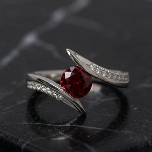 May include: A silver ring with a large, round, deep red gemstone at the center. The band splits into two curving arms, each lined with small, sparkling white stones. The ring is set against a dark, marbled surface.