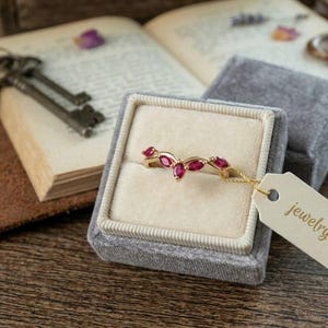 May include: A gold ring with marquise-cut ruby gemstones, displayed in a gray velvet ring box. The ring is on a tag that reads "jewelryinspireco". The background includes an open book, keys, and a leather surface.