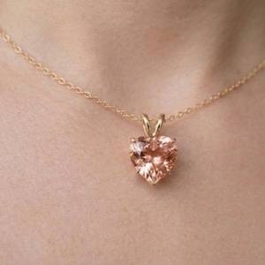 May include: A gold necklace featuring a heart-shaped pendant. The pendant is a peach-coloured gemstone, and the chain is delicate. The necklace is worn around the neck.
