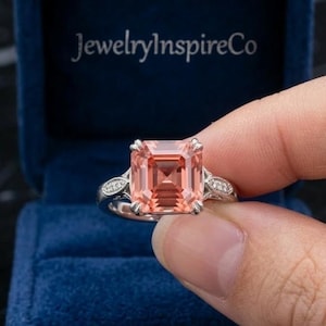 May include: A silver ring with a large, rectangular, peach-colored gemstone is held in a blue velvet ring box. The ring has a detailed band with small accent stones. The JewelryInspireCo logo is visible.
