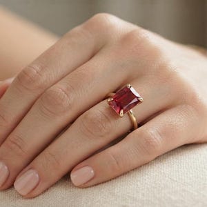 May include: A gold ring featuring a large, rectangular, emerald-cut deep red gemstone set in a four-prong setting. The band is a simple, polished gold metal. The gemstone displays a vibrant, saturated crimson hue with clear facets that reflect light.
