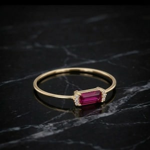 May include: A gold ring with a rectangular, deep pink gemstone at the center, flanked by small, clear accent stones. The ring band is thin and polished, set against a dark, marbled surface.