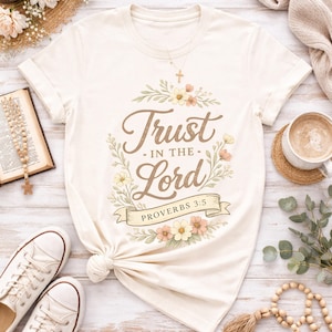 May include: Cream-colored t-shirt featuring the text "Trust in the Lord" in elegant script, surrounded by a floral wreath. The design includes a banner with "Proverbs 3:5." The shirt is styled with rolled sleeves and a knotted hem.