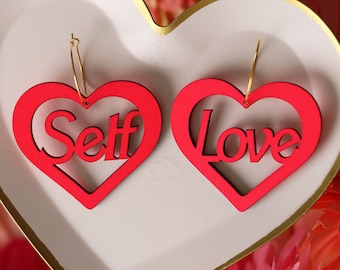 Op de afbeelding: Een paar rode hartvormige oorbellen met de woorden "Self Love" uitgesneden. De oorbellen worden gepresenteerd op een wit hartvormig schaaltje.