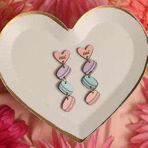 Puede incluir: Un par de pendientes de madera con un dije de corazón rosa y tres dijes de macarons en rosa, azul y morado. Los dijes están apilados uno encima del otro y el dije de corazón está en la parte superior. Los pendientes están en un plato blanco en forma de corazón. Los dijes de corazón tienen la palabra "oui" escrita en ellos.