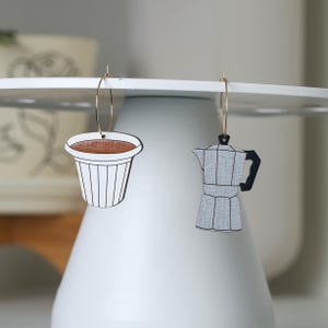 May include: A pair of gold hoop earrings with a coffee cup and a coffee pot charm. The coffee cup charm is white with a brown coffee inside and black lines. The coffee pot charm is gray with a black handle and spout.