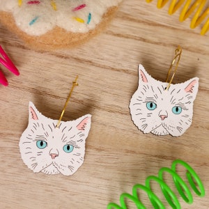 May include: A pair of cat face earrings with gold hoops. The cat faces are white with blue eyes and black whiskers.