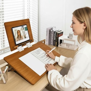 Könnte beinhalten: Ein hölzerner Schreibtisch-Organizer mit verstellbarem Tablethalter und Schreibfläche. Das Tablet zeigt einen Videoanruf an. Eine Person schreibt mit einem Stift auf einen Notizblock. Der Schreibtisch ist aus Holz.