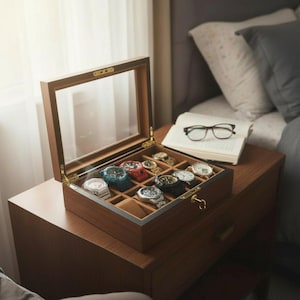 Może przedstawiać: Drewniane pudełko na zegarki ze szklaną pokrywą, prezentujące kolekcję zegarków na rękę. Pudełko jest otwarte, odsłaniając przegródki wyłożone miękkim, brązowym materiałem. Zegarki mają paski i tarcze w różnych kolorach.