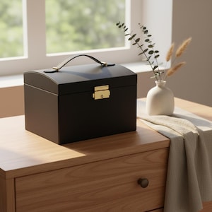 Pode incluir: Uma caixa de joias preta com uma parte superior curva e uma alça. A caixa tem um fecho e acessórios de alça dourados. A caixa está em uma superfície de madeira.