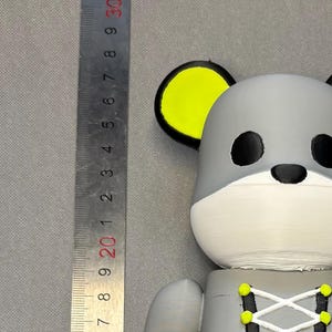 May include: A gray and black bear-shaped figurine with yellow accents. The figurine has a white muzzle, black eyes, and yellow ears and hands. The bear is wearing a corset-style top with white lacing. The figurine is approximately 10 inches tall.