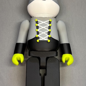 May include: A gray and black figurine with a bear-like head and a corset-style design. The figurine has yellow accents on the ears, hands, and feet. The corset is laced with white string and yellow eyelets. The overall design is modern and playful.