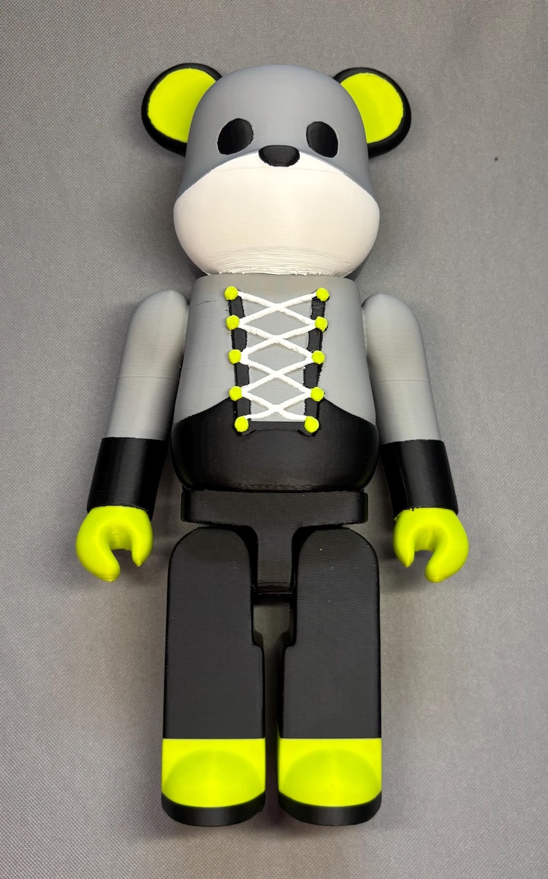 May include: A gray and black figurine with a bear-like head and a corset-style design. The figurine has yellow accents on the ears, hands, and feet. The corset is laced with white string and yellow eyelets. The overall design is modern and playful.