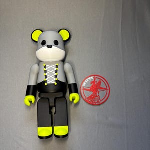 May include: A gray and black bear-shaped figurine with yellow accents on the ears, hands, and feet. The figurine has a white face and a laced-up design on the chest. A red and gray circular emblem with the word "GERRIES" is next to the figurine.