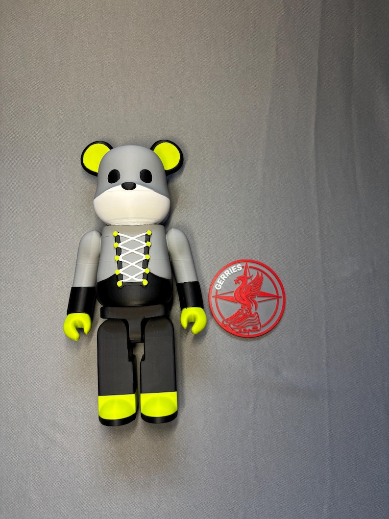 May include: A gray and black bear-shaped figurine with yellow accents on the ears, hands, and feet. The figurine has a white face and a laced-up design on the chest. A red and gray circular emblem with the word "GERRIES" is next to the figurine.