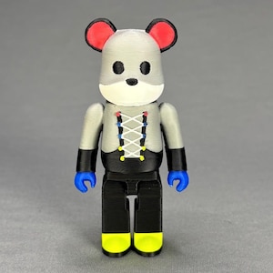 Puede incluir: Una figura impresa en 3D con forma de rat&oacute;n. La figura tiene una cabeza gris con orejas rojas, ojos negros y un hocico blanco. Viste una parte superior gris con un dise&ntilde;o de cors&eacute; negro y pantalones negros. Las manos son azules y los pies amarillos.