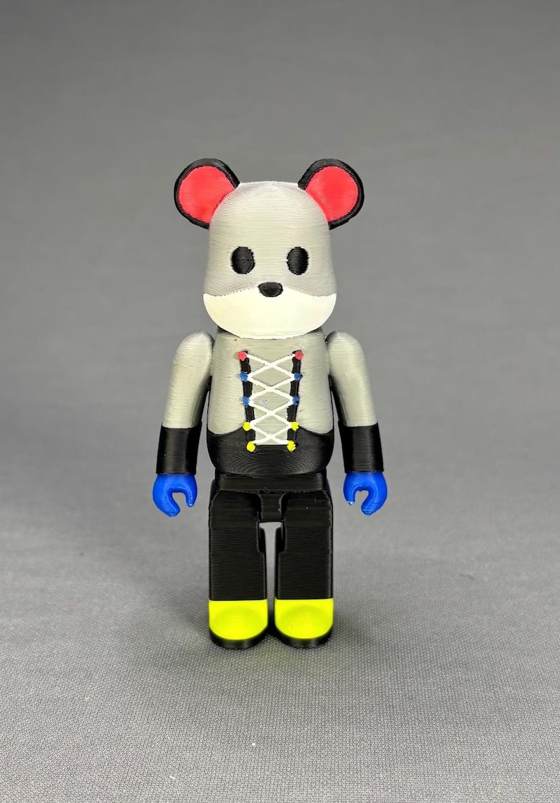 Puede incluir: Una figura impresa en 3D con forma de rat&oacute;n. La figura tiene una cabeza gris con orejas rojas, ojos negros y un hocico blanco. Viste una parte superior gris con un dise&ntilde;o de cors&eacute; negro y pantalones negros. Las manos son azules y los pies amarillos.