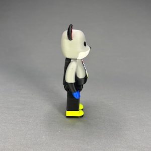 Puede incluir: Una figura gris con forma de oso con brazos y piernas negros, manos azules y pies amarillos. El oso tiene un hocico blanco, ojos negros y detalles rojos en las orejas. Lleva un atuendo negro con detalles coloridos.