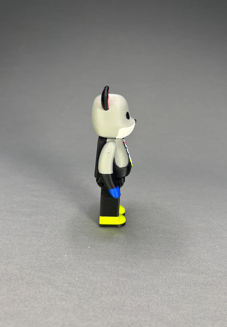 Puede incluir: Una figura gris con forma de oso con brazos y piernas negros, manos azules y pies amarillos. El oso tiene un hocico blanco, ojos negros y detalles rojos en las orejas. Lleva un atuendo negro con detalles coloridos.