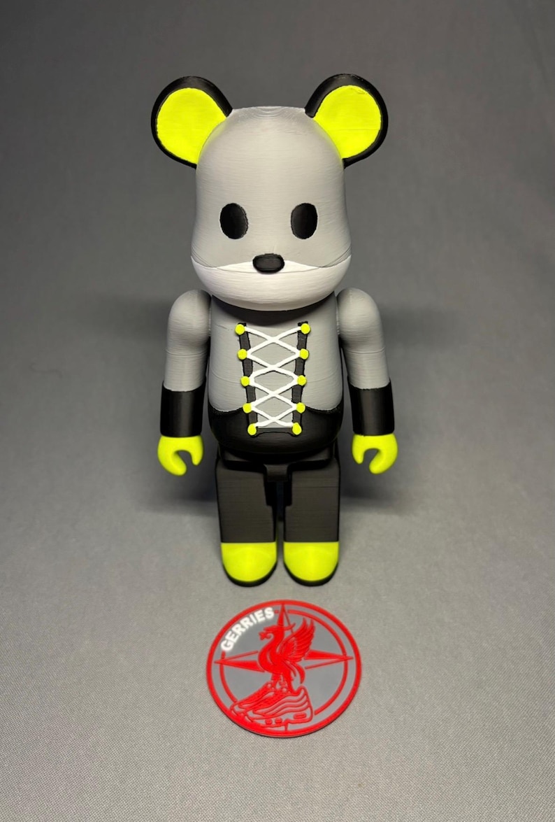 May include: A gray and black mouse-shaped figurine with yellow accents on the ears, hands, and feet. The figurine has a white laced-up design on the chest and black eyes. A red and gray logo with the word "GERRIES" is at the base.