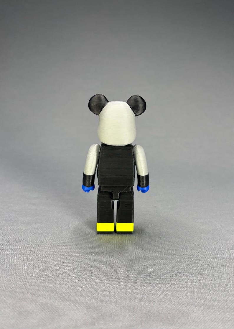 Puede incluir: Figura impresa en 3D con tem&aacute;tica de panda. La figura tiene una cabeza blanca con orejas negras, un torso negro y piernas negras con pies amarillos. Los brazos son plateados y las manos azules. Dise&ntilde;o estilizado y en bloques.