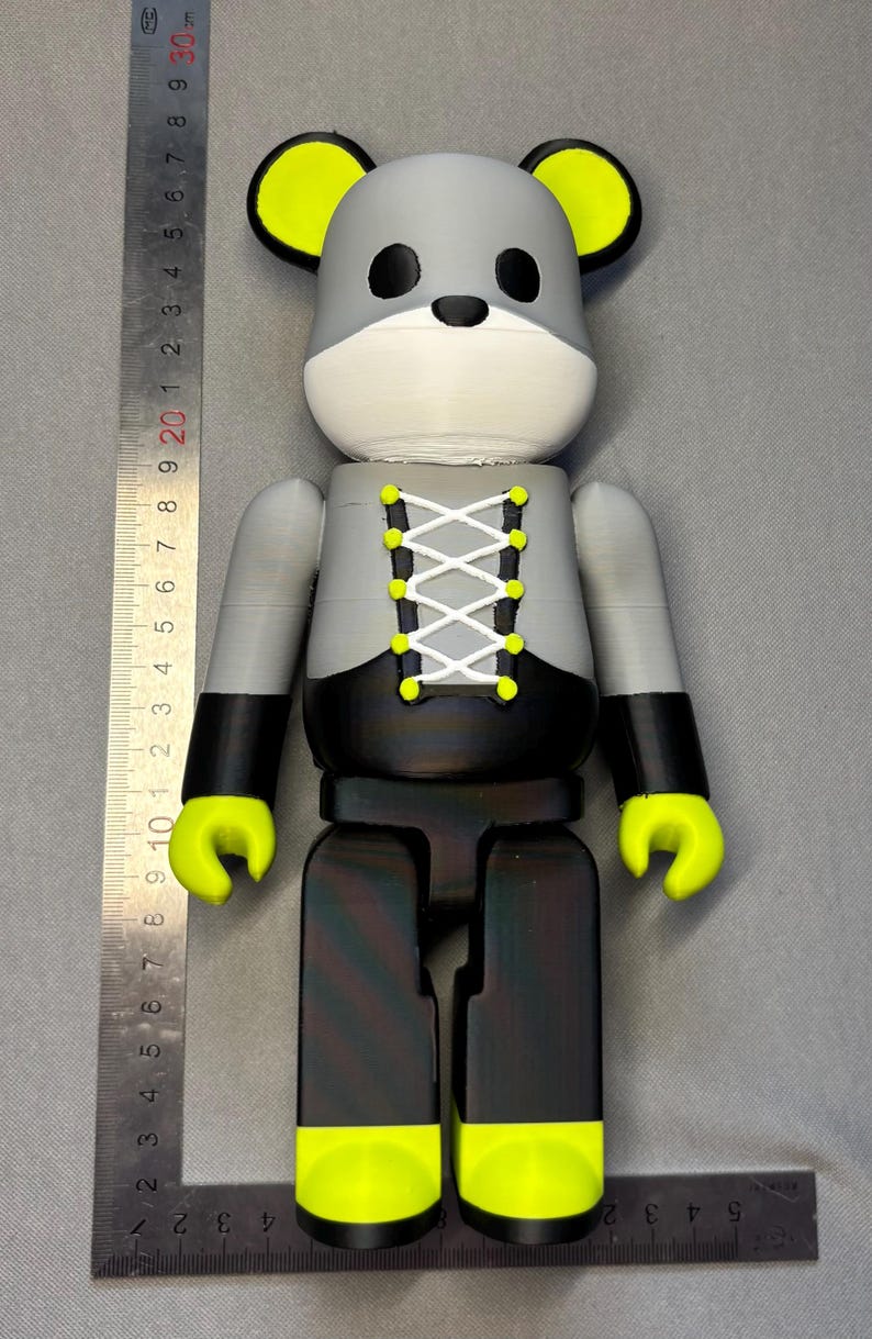 May include: A gray and black bear-shaped figurine with yellow accents. The figurine has a white muzzle, black eyes, and yellow ears and hands. The bear is wearing a corset-style top with white lacing. The figurine is approximately 10 inches tall.