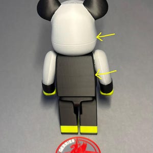 May include: A stylized bear-shaped figurine in white, black, and yellow. The figure has a white head with black ears, a black torso, and black legs with yellow accents. A red and gray logo is at the base.