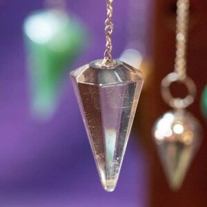 May include: Close-up of a clear crystal pendulum with a silver chain, hanging in front of a blurred purple background. Other pendulums with green and silver accents are also visible, suggesting a spiritual or metaphysical theme.