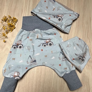 Puede incluir: Un conjunto de ropa de bebé con pantalones azul claro, un gorro a juego y un calentador de cuello. El conjunto está estampado con tractores marrones, árboles y puntos rojos. Los pantalones tienen una cintura y puños a rayas azul oscuro.
